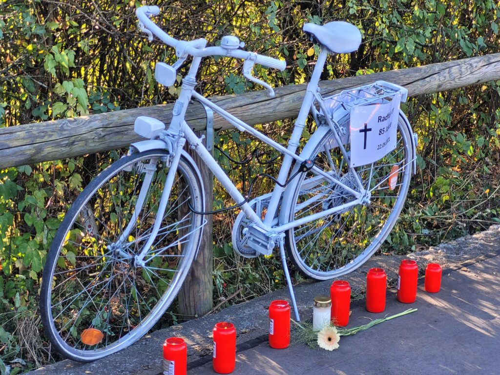 Foto eines Ghostbikes, das zum Gedenken an einen getöteten Radfahrer aufgestellt wurde. Es sind Kerzen und Blumen davor.